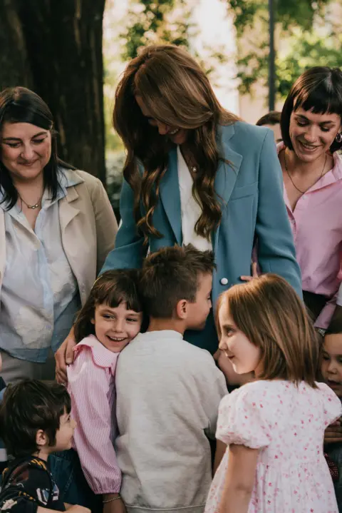 Royal Foundation A smiling Kate in blue suit is embraced by small children, surrounded by smiling women