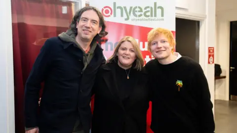 Bradley Quinn Gary Lightbody and Ed Sheeran at the Oh Yeah Music centre. Gary wears a khaki hoody, with a navy jacket over it and jeans. He has curly longish hair. Ed Sheeran has shaggy red hair and wears a black jumper with a colourful pineapple logo on it. They are standing on either side of a woman in a long black coat, black jumper and acorn necklace.