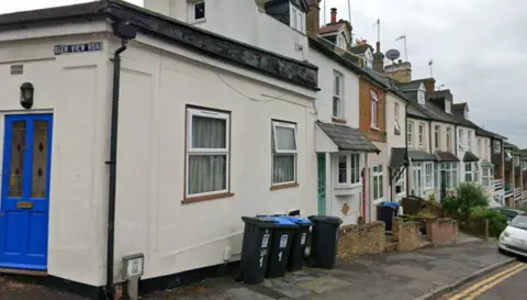 Google A row of houses on a street which is on a slight hill. There are bins outside the houses and there is a white car parked half on the pavement. There is a sign on the corner saying "Glen View Road"