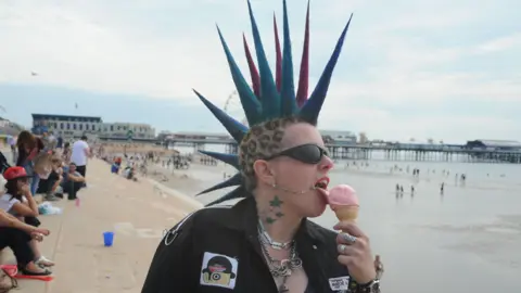 A punk with multicoloured spiked hair licks an ice cream on the Blackpool promenade. A pier is in the background.