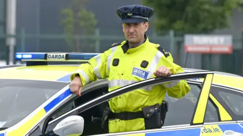 An Garda Síochána A man wearing an Irish police officer uniform of a yellow hi-vis jacket standing behind the open door of a police car which is stationary