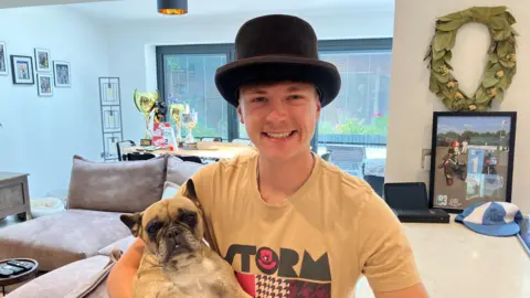 A 23-year-old young man showing off a nice big smile, wearing his top hat and a beige T-shirt with the word 'storm' on the front. He is sitting in the family kitchen holding his dog Dolly on his knee, which is a creamy, beige French Bulldog. Behind there is a picture of him in a frame on the breakfast bar, when he came first at the Goodwood Motorsport event in April. There is also a selection of big trophies and medals on the table in the background.