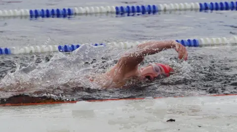 Erin Medcalf Woman (Erin Medcalf) mid-swim. You van see her wearing a bright red swim hat and transparent goggle. Behind her in the background, there is a white and blue land divider.