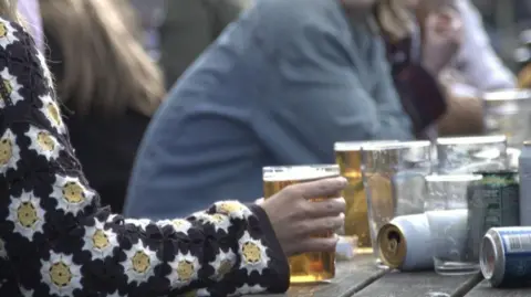 A woman's arm rests on a wooden table as she holds a pint of beer. Other people are sat at the table which is has a number of other drinks cans and glasses on it, some of which are empty