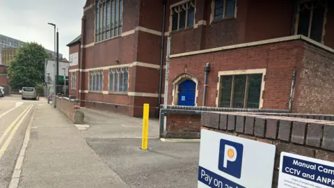 Emma Baugh/BBC Image of Geneva Street, where there is a red-brick building and signs for a car park, it has double yellow lines on the road.