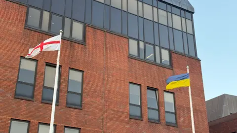 The st george's cross flying on flag pole on the left next to the ukrainian flag on the right outside a red bricked office buidling.