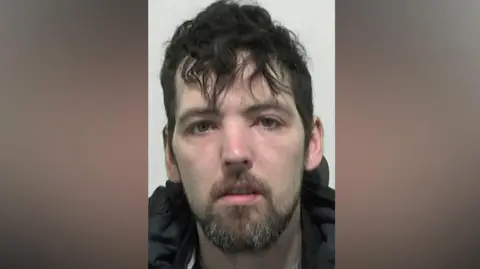 Mugshot of Hepplewhite. He has scruffy black hair and a shore dark beard.