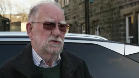 An older man with a white beard and wearing dark lens glasses stands in front of a car, itself next to what looks like a cottage built in Yorkshire stone.