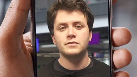 A man with brunette hair is looking towards the camera and is in a phone held by a hand.