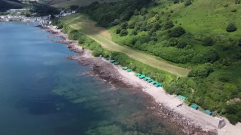 A drone shot of a row of about 15 green temporary huts on a rocky beach the sea is green and clear below them and behind them are green fields. Further up the coast you can see a village.