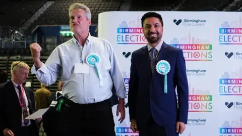PA Two men, one in a light blue shirt and dark trousers, the other in a dark blue suit, stand next to each other. Both are wearing pale blue rosettes. One has clenched his fist in a celebratory way and is looking off camera. the other is smiling into the camera. A big banner with birmingham elections written on it is behind them.