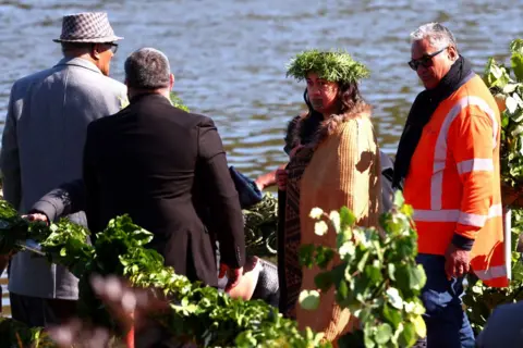 Getty Images Maori Queen Kuini Nga wai hono i te po stands next to a river wearing a headress and a cloak, with three men standing alongside her.