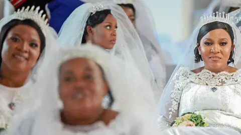 Several brides, dressed in white wedding gowns, veils and tiaras - one holding a bouquet - wait for the mass wedding to get under way