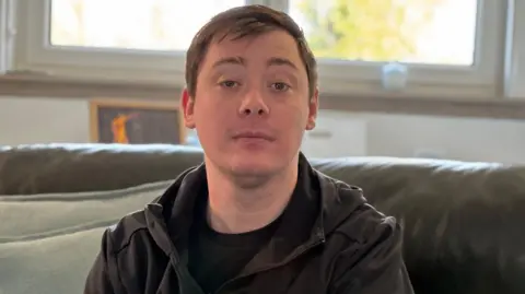 Stephen Reside, who has brown hair combed in a side shed, sits on a leather couch in a living room. he is looking directly at the camera with a serious expression on his face.
