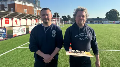 Two man in dark clothing stood on a football pitch. They are facing the camera, the man on the right is holding a wooden board with oysters on