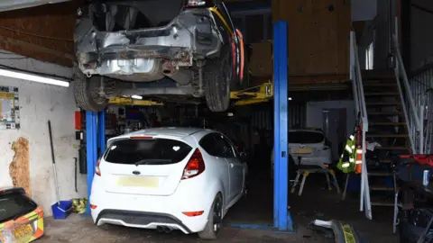 Essex Police A white car on the ground and a black car on a platform above it in a lock-up. The black car has been partially stripped of parts.