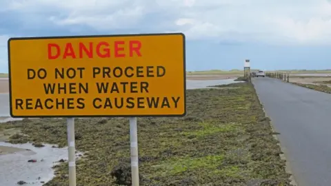 An orange rectangular sign has the word 'danger' written in red and the words 'do not proceed when water reaches causeway' below in black. Next to it is a road crossing a large stretch of sand over a bridge with an island in the distance.