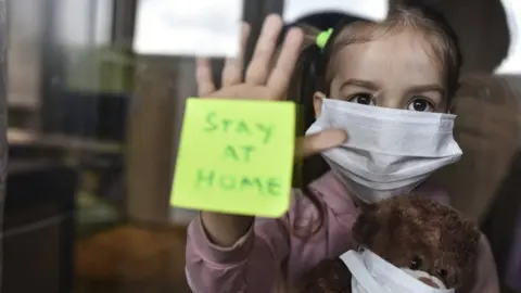 Getty Images Young girl with face covering holding a teddy bear with face covering holds her hand against a window, with a note stuck to the window reading "Stay at home"