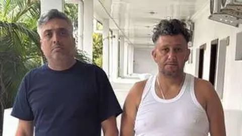 Carlos Alberto Rodelo, in blue T-shirt and shorts, stands next to Jorge Cubillos, in white vest and black trousers, with hands in pockets, on the balcony of their small hotel room in Kinshasa, with palm trees in the background.