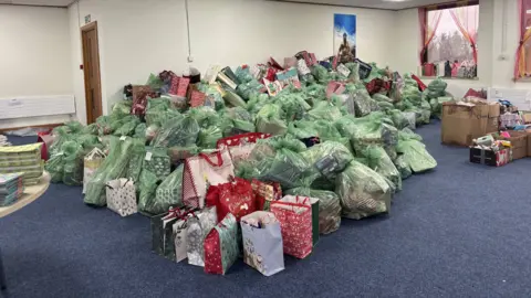 A huge pile of wrapped Christmas presents that are inside green plastic bags. The bags are in a giant mound stretching across a large room