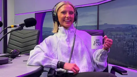 Alysia Mcandrew Alysia Mcandrew in the BBC Radio Lancashire studio. She has short, straight blonde hair, and is wearing a white running jacket and over-ear headphones. She is holding a BBC branded mug and smiles at the camera.