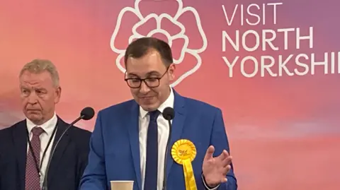 Harrogate and Knaresborough MP Tom Gordon gives his acceptance speech after becoming MP in July 2024. He is standing behind a podium at the count centre, wearing a blue suit and white shirt with dark blue tie. He has glasses on and wears a yellow Liberal Democrat rosette.