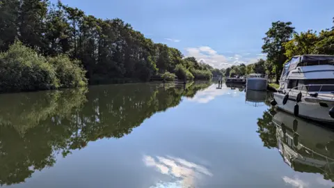 Peter Akrill TUESDAY - A beautiful sunny day at Abingdon Lock with the sun shining on the waterway. There are green trees overhanging the left bank and a white yacht moored on the right bank. The sun and small white clouds are reflected in the still water