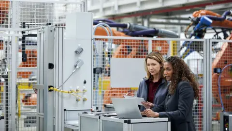 Morsa Images / Getty Images Female workers in a car factory discussing technological solutions