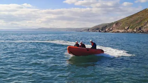  A picture of lifeguards on a lifeboat travelling across a stretch of water. The boat is orange in colour and there is three people sat on it. There is a large rock face in the distance.