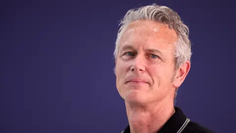 Getty Images Mark Foster stands against a deep purple background wearing a black polo shirt. He is smiling and looking directly at the camera