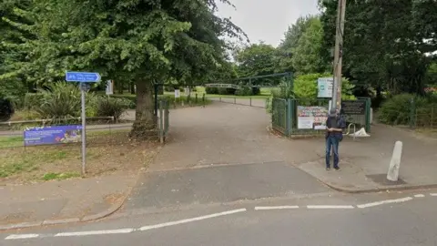 Google Entrance to a park from a road with green gates either side and a metal green barrier across the top to restrict vehicle height. A man stands to the right of the entrance on the pavement, looking at his phone. There are signs either side of the entrance