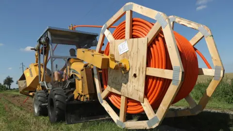 Getty Images A large work vehicle carrying a mammoth spool of reel rides across open green grassland