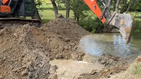 Ellen Knight/BBC A large orange digger perched on an earth bank, with the scoop pointing downwards towards the water. Water rushes through where the digger has lifted out soil, with the water starting to pool in places as it flows into the new channel. The banks are lined with trees and shrubbery. 