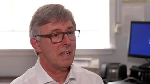 Dr John Montgomery sitting in an office with a computer screen in the background. He is wearing a white shirt and has glasses.