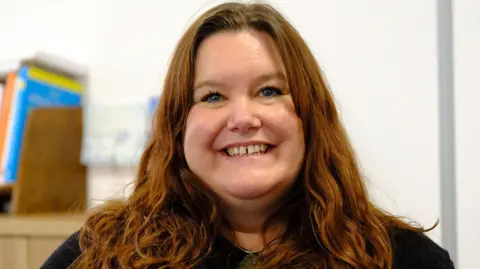 Shetland Islands Council A woman with long wavy auburn hair wearing a dark textured top, seated indoors with a blurred background featuring shelves and colourful books.