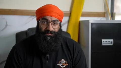 Un hombre con una barba negra muy poblada, bigote, gafas plateadas de montura fina y un turbante naranja. Lleva una chaqueta negra con un emblema que dice Ejército Sikh.