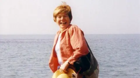 Claire Ashton Woman wearing orange shirt, with bag over one shoulder, holding a hat, while smile on the beach, with the sea behind her