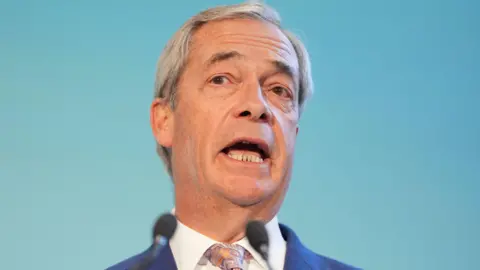 Nigel Farage wearing a blue suit jacket, white shirt, and a patterned tie, standing in front of a light blue background with two microphones positioned in front.”