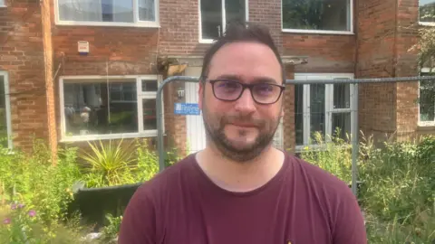 Christian Wakeford, the Labour MP for Bury South, is pictured in front of the vandalised apartment block on Baguley Crescent. He is wearing a claret t-shirt and black glasses. He has a short dark beard and short hair. Behind him, the building is surrounded by overgrown plants and there is a single moveable grey metal wire security fence panel.