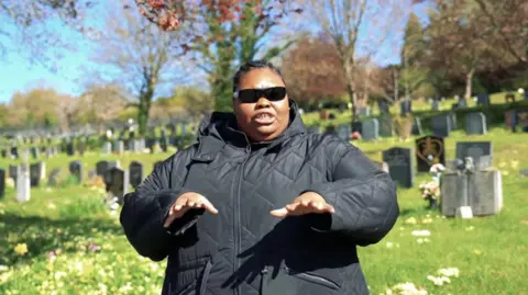 Wycombe Youth Action / Love Music Hate Violence A person wearing a black quilted jacket and dark sunglasses stands in the foreground of a cemetery, with rows of gravestones stretching across a gently sloping grassy landscape behind. The person has both hands held out in front at waist height. Trees with sparse spring foliage frame the scene, and clusters of flowers are visible among the graves. Bright sunlight illuminates the grass and headstones beneath a clear blue sky, creating strong contrast between the sunlit background and the dark clothing in the foreground.