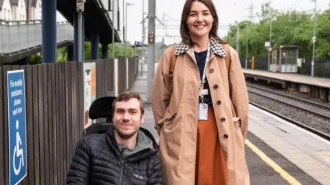 A man in a wheelchair and a woman stand on a train platform. A sign next to the man indicates a waiting area for mobility assistance.
