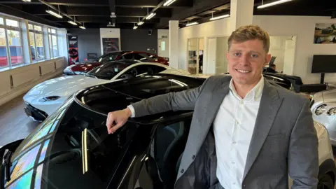 Martin Heath/BBC Will Chapman with short light brown hair wearing a grey jacket and white shirt, smiling at the camera and leaning on a car. There are other cars parked beyond it in a car showroom with windows to the right and white-walled offices to the left.