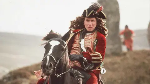 Getty Images Tim Roth in powdered wig and red coat rides a horse in Rob Roy, an action shot with a hill behind him and other soldiers on the grass.