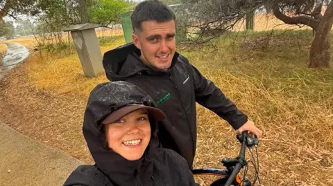 Cai James Annie and Cai standing next to a bike, wearing black rain coats. They are both smiling whilst looking at the camera.