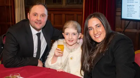Pete Hill Photography Heidi and her parents seated closely together at a round table covered with a deep red tablecloth. Heidi is in the middle holding a glass filled with what appears to be orange juice.
On the table in front of them are several items: two glasses containing an orange beverage, a glass of what appears to be a sparkling drink, a gold glittery bag, a light pink phone case, and a white plush toy lying near the table’s edge. A red curtain hangs behind the group, and a large screen displaying a list of names is visible on the right side of the image.