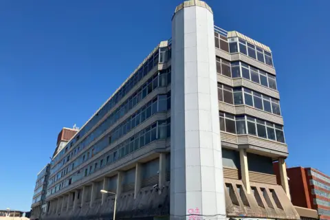 Owen Sennitt/BBC Sovereign House, taken from the southern side up close, looking upwards, showing a cylindrical tower connected to the side of the concrete office block