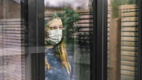 Getty Images Woman stands behind window wearing face mask