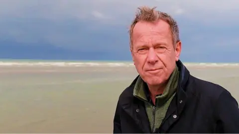 A man, BBC correspondent Andrew Harding, stands in front of the sea