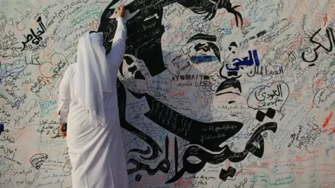 Reuters A man writes on a painting depicting Qatar's Emir Sheikh Tamim Bin Hamad Al Thani in Doha, Qatar (2 July 2017)