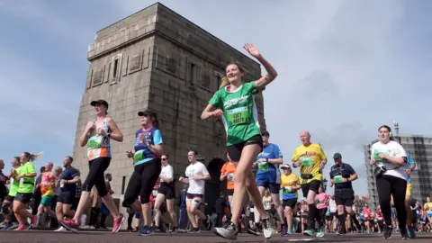 PA Media Woman in green Macmillan cancer support shirt waves to someone off camera as she takes part in the run